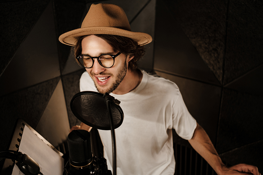 Portrait of attractive stylish male musician confidently singing in microphone recording song in professional sound studio